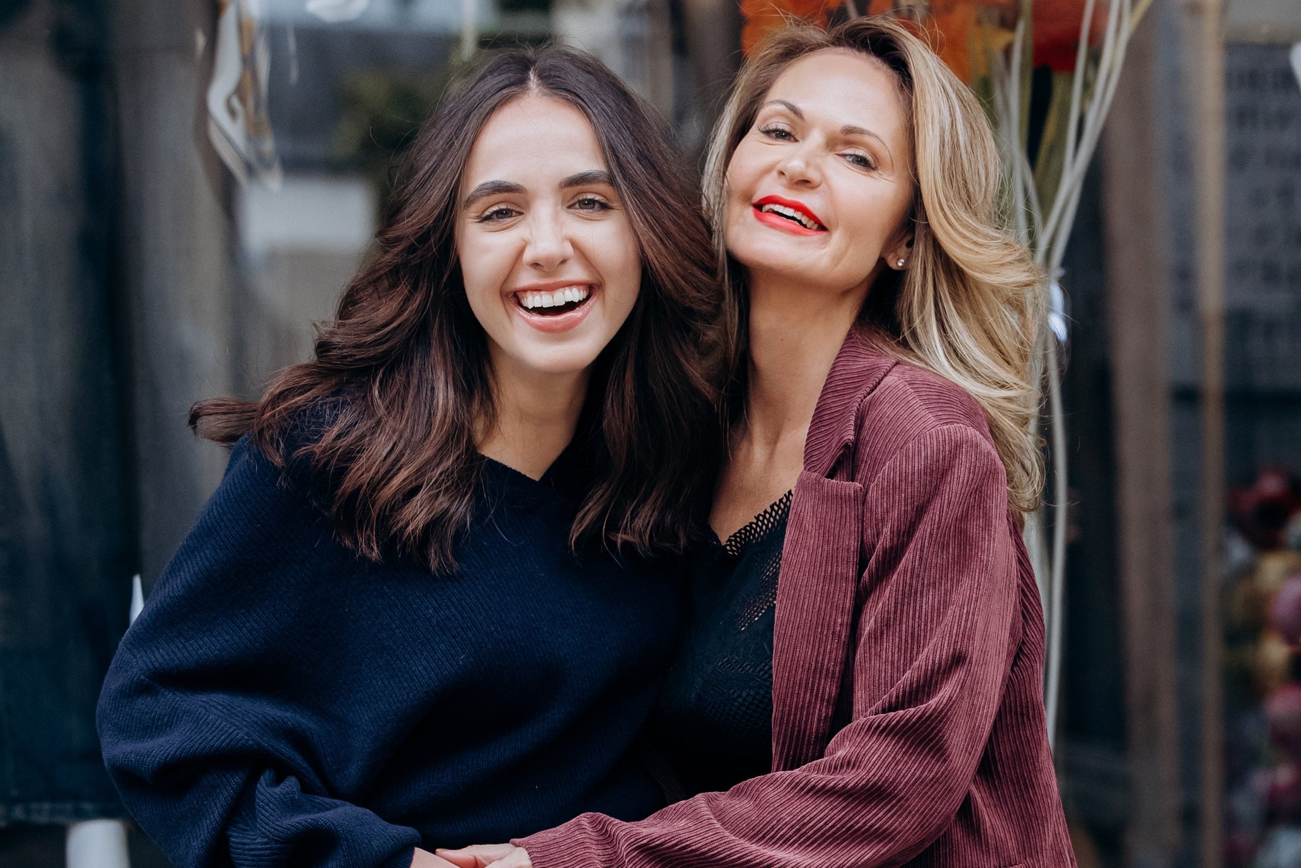 Two women standing together in front of a store window with various items displayed.